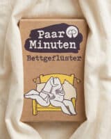 Schachtel PaarMinuten Bettgeflüster auf weißer Bettwäsche, auf der Verpackung ein illustriertes Paar in einem gelben Bett