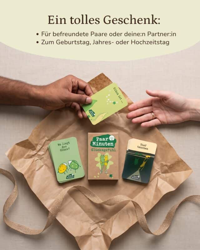 Zwei Hände reichen einander eine hellgrüne Karte „Glück ist …“ über Kraftpapier; darunter liegen die Schachtel PaarMinuten Glücksgefühl und zwei Kartenstapel der Kategorien „Wo liegt das Glück?“ und „Tief tauchen“