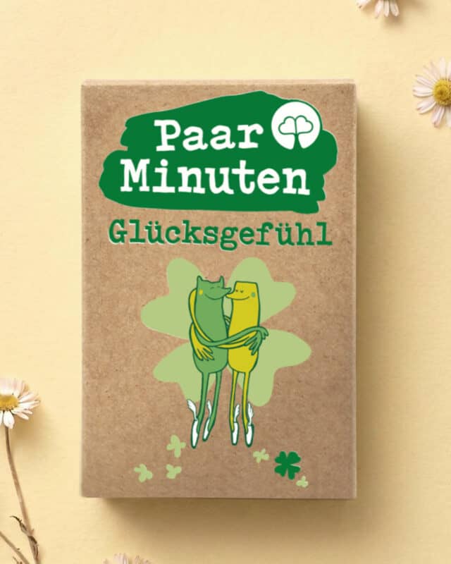 Schachtel PaarMinuten Glücksgefühl auf gelbem Untergrund mit Gänseblümchen, darauf zwei sich umarmende Figuren vor einem Kleeblatt