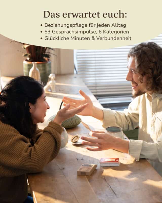 Frau und Mann sitzen sich an einem sonnigen Holztisch gegenüber, lachen und strecken die Hände zueinander aus; vor ihnen liegen die Kraftpapier-Schachtel und ein Kartenstapel