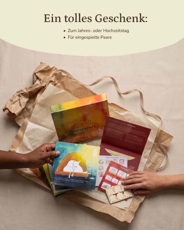 Die aquarellierte Sofasafari-Box liegt mit aufgefächerten Erlebnisumschlägen, einem aufgeklappten roten Umschlag, Stickerbogen und Holzwürfel auf Kraftpapier; zwei Hände greifen von links und rechts ins Bild